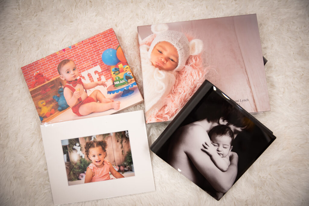 Picture of acrylic photo album with baby on it. A Canvas print with one year old baby in red. A 5x7 print with a toddler girl smiling. And a photo album box with newborn girl in white. All examples of printing photographs. 