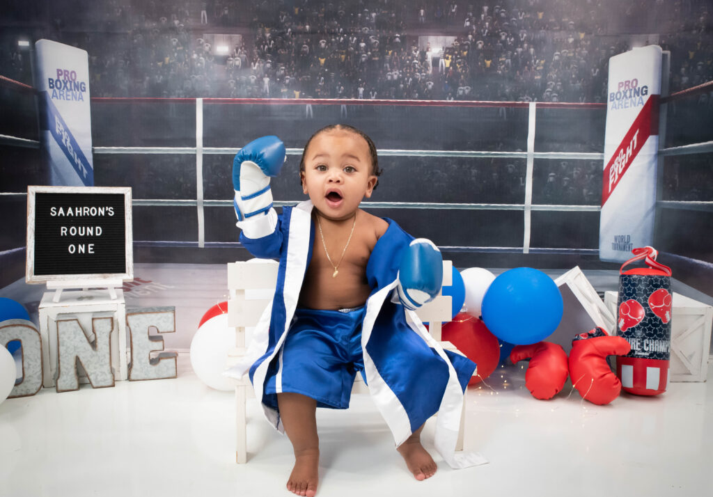 Baby Boy in boxing ring with blue robe and boxing gloves celebrating his first birthday with a themed cake smash by Neptune, New Jersey Photographer. 